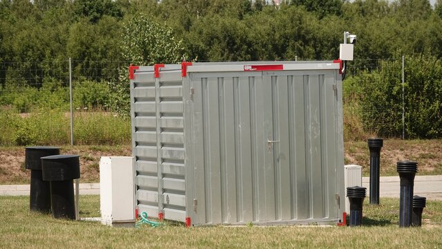 Techniczny, metalowy kontener z blachy falistej stojący na trawie pod lasem. Obiekt wyposażony jest w kamerę, a wok&oacute;ł niego rozmieszczone są czarne rury i białe skrzynki instalacyjne.