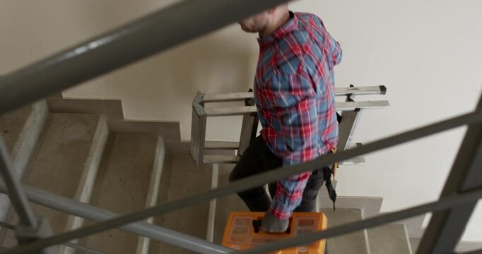 A man carries a toolbox and a compact ladder as he climbs the stairs between floors. A plumber is called to service the utilities in an apartment building.