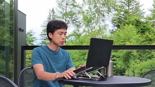 Young man focused on laptop at outdoor table. Laptop rests on stand, angled for comfort. Green trees frame background, bright daylight. Natural setting, quiet, productive, relaxed vibe