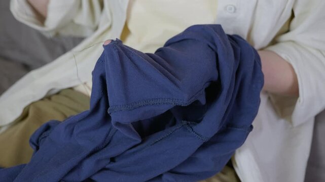 Woman's hands mending blue clothes with needle and thread. Close up view of a woman's hands using a needle, thread, and a thimble to carefully mend a hole in a blue garment