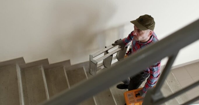 A man wearing a baseball cap climbs the stairs of a multi-story building while holding a portable ladder and a toolbox.