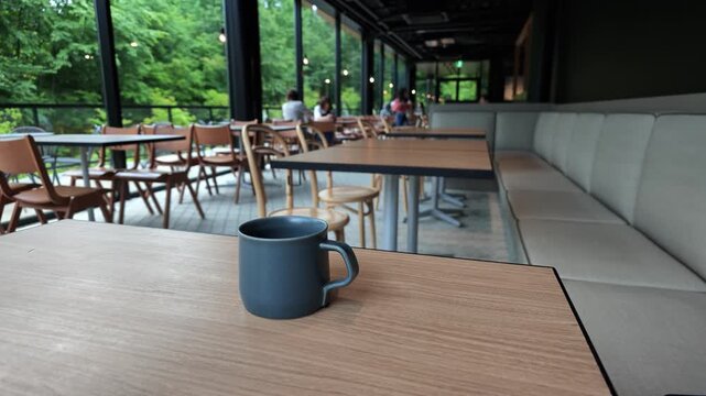 Calm coffee cup sits on wooden table. Large windows frame green forest view. Modern cafe interior with cozy seating. People enjoy quiet moments nearby. Ideal for travel, relaxation