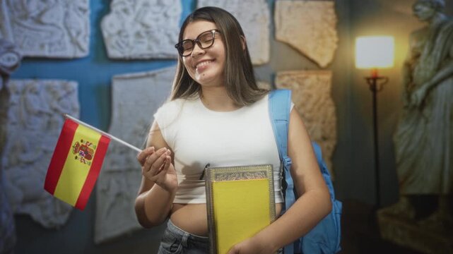 Young hispanic woman holding spanish flag and books, smiling with glasses and backpack while holding the flagpole in a building; study abroad pride.