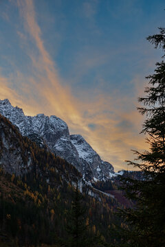 Alpine landscape with snow-covered mountain peaks and autumn coloured forested hills stands against golden coloured clouds during dramatic sunset. Located in the Gosau Valley in Upper Austria.