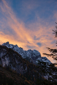 The alpine landscape with snowy towering mountain peaks against dramatic clouds glowing with pink and yellow hues during sunset. Located in the Gosau Valley in Upper Austria.