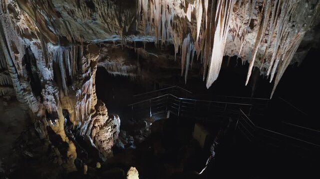 Limestone cave chamber with dripping water, dramatic stalactites, and lit walkway, suited for geotourism, karst geology, subterranean exploration, and underground hydrology themes.