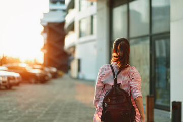 A woman with a backpack walks away along an urban street at sunset, warm light framing her...