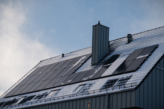 House roof equipped with photovoltaic solar panels partially covered in snow on a bright winter day. 