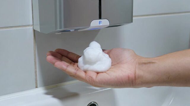 Man placing hand under automatic foam soap dispenser in restroom. Hygiene process with contactless device in bathroom. Sanitization and health care routine concept