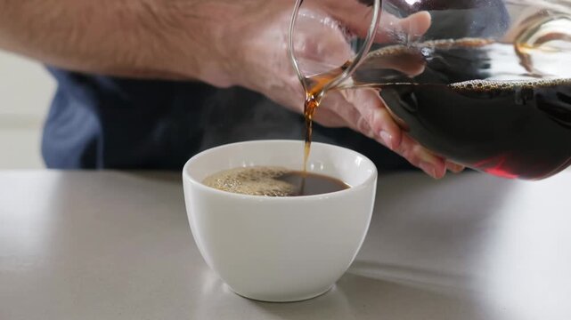 Man pouring hot black coffee from glass carafe into white ceramic cup. Close up view of morning beverage preparation. Steam rising from fresh drink in kitchen