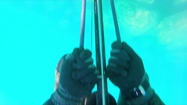 First person point of view of a spearfisherman wearing gloves loading a speargun with rubber bands in clear turquoise water over a rocky seabed, preparing for spearfishing