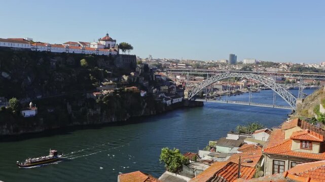 Sunny day in porto with the iconic luis i bridge and vila nova de gaia on the horizon as a traditional rabelo boat sails along the douro river, passing the historic ribeira district