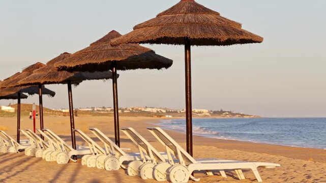 Deserted sandy beach featuring a row of empty sun loungers and straw parasols with gentle ocean waves in background, creating a serene and tranquil atmosphere at the end of the day. Algarve, Portugal