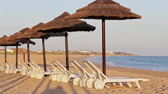 Tranquil scene showing a row of empty sunbeds and thatched umbrellas on a beautiful sandy beach during a calm, golden sunset, with gentle ocean waves in the background. Algarve, Portugal