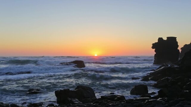 Magnificent ocean waves crashing on a rugged coastline during a beautiful sunset, with the sun dipping below the horizon and casting a warm, golden light across the tranquil evening sky