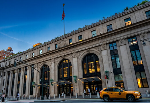 New York, NY - US - Jul 20, 2026 Evening view of Moynihan Train Hall in New York City, its Beaux-Arts facade glowing with warm interior light as a yellow taxi passes along a busy Manhattan street.