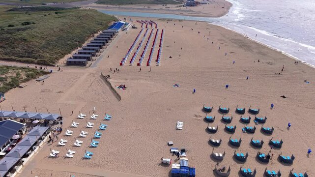 Beautiful high altitude aerial panoramic perspective of organized beach resort with pavilion dining terrace sunbeds hooded windbreak pods and cabins along ocean coast under bright sunny sky
