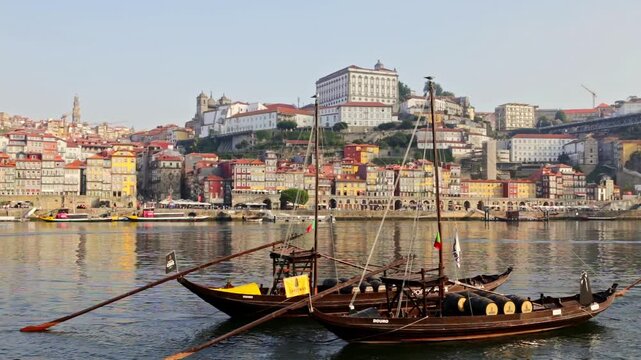 Traditional rabelo boats carrying wine barrels gently floating on the douro river with the colorful and historic ribeira district of porto, portugal, visible in the sunny background