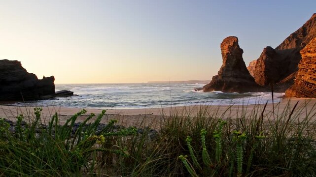 Secluded sandy beach along the vicentina coast in portugal, with towering rock formations and cliffs illuminated by the warm light of a beautiful sunset as ocean waves gently wash ashore
