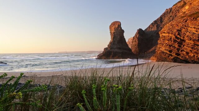 Dramatic sea stacks and cliffs on a sandy beach basking in the warm, golden light of sunset, with gentle waves from the atlantic ocean washing ashore on the beautiful vicentina coast