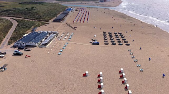 Beautiful high altitude aerial panoramic perspective of organized sandy beach resort with pavilion lounge chairs windbreaks and beach cabins alongside rolling ocean waves under bright sunlight