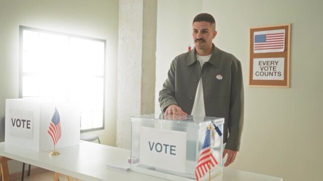 Young hispanic man places ballot into a clear ballot box while wearing an i voted sticker inside a polling building; civic pride duty.