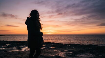 Fototapeta premium Silhouette of a woman standing at sunset on rocky coastline, wind blowing hair, warm golden light reflecting on calm waves, serene atmosphere captured from low angle.