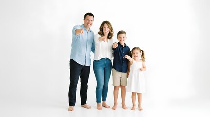 Naklejka premium A happy family of four stands together in a white studio, the parents and two children all pointing forward with smiles, barefoot and dressed casually in blue and white clothing, looking directly at