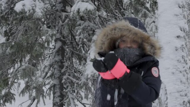 Girl in a dark hooded parka with fur trim and pink gloves stands in a snowy spruce forest, adjusting her hood in the cold