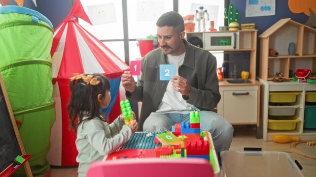 Man holding number cards and showing them to a girl at a play table with colorful blocks and tent in a building; early learning nurturing.
