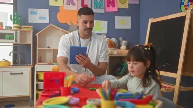 Male teacher holds tablet and offers toy while watching a girl arrange colorful play blocks at a preschool classroom table with chalkboard and toys; learning calm.
