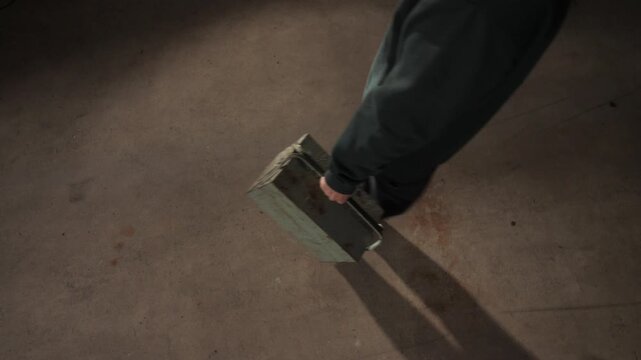 High angle view person walking while carrying an old green metal toolbox with a handle, casting a shadow on a textured concrete floor in an industrial or workshop setting
