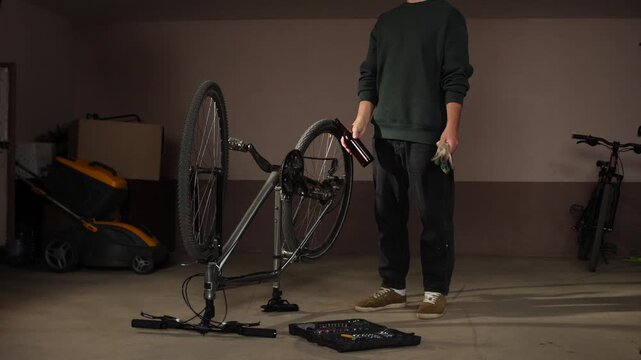 Man standing in a garage, holding a bottle and work gloves, while working on an upturned bicycle with a tools case on the floor, focusing on maintenance