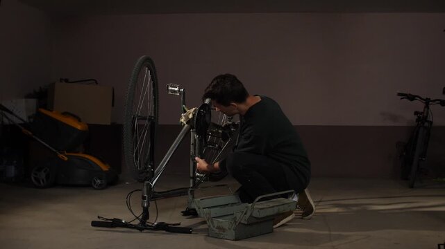 Man in a dark jersey crouching near a bicycle with its wheel off, carefully working on the bike's chain and gears, using tools from a metal toolbox on the concrete floor of a home garage