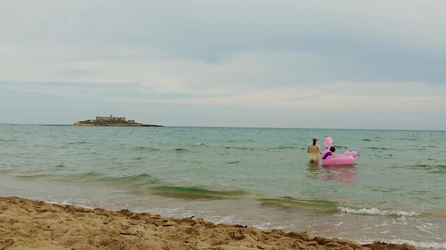 Mother and toddler enjoying a summer afternoon by the Mediterranean Sea with a pink flamingo float and Isola delle Correnti in the background.