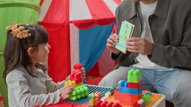 Girl stacking colorful plastic blocks while adult reads picture flashcards and smiles in an indoor playroom building; curiosity learning.