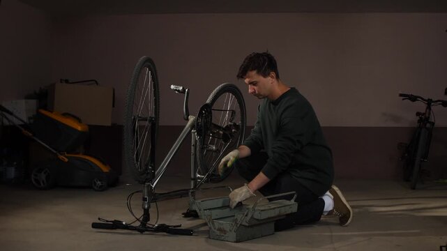 Man kneeling on the floor, wearing working gloves and opening a metallic toolbox while working on a bicycle that is positioned upside down in a home garage, performing maintenance