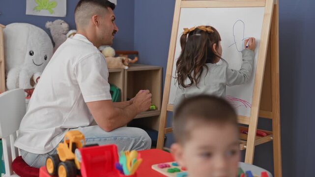 Man teacher kneels beside girl drawing on an easel, hand holding marker guiding strokes in studio; nurturing learning bond.