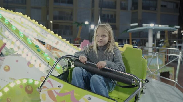 Young girl with long blonde hair smiles while riding a brightly lit amusement park ride at night at a seaside funfair