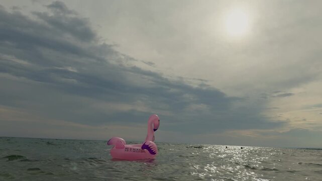 Minimalist composition featuring a solitary flamingo float and swimmers in the background, illuminated by a soft backlight sun over calm Mediterranean waters.