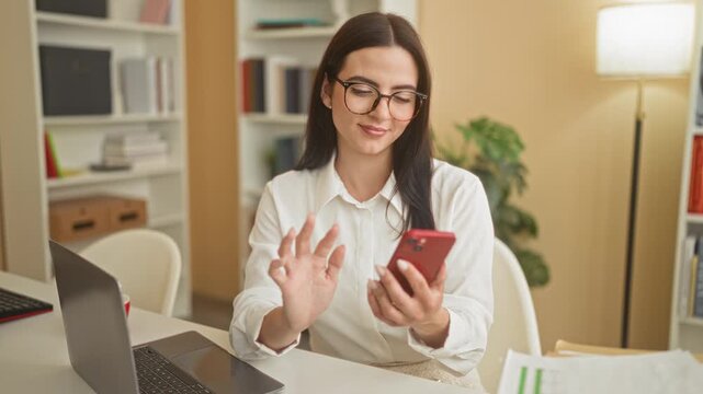 Young hispanic woman tapping smartphone with fingers while seated at laptop desk in building office, papers and lamp visible; focused productivity.