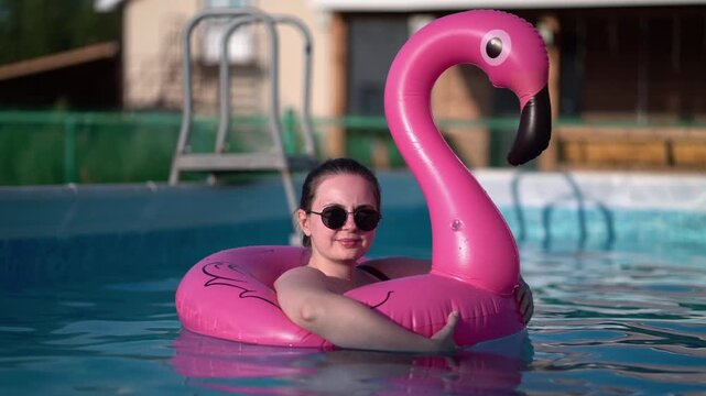 Young woman in sunglasses relaxes smiling in a pink flamingo inflatable float in a sunny outdoor swimming pool on a hot summer day at a resort.