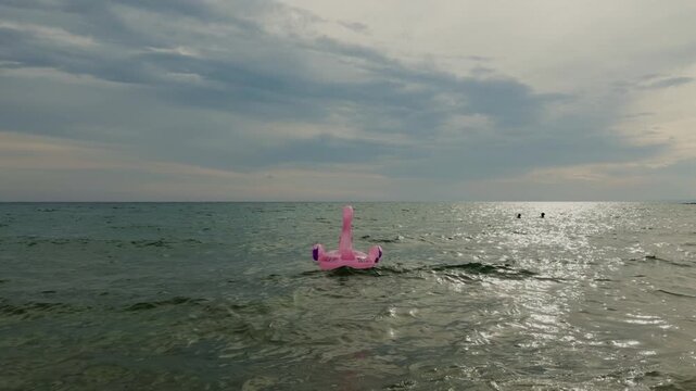 Backlit pink flamingo float drifting across sparkling Mediterranean water, with swimmers in the background and a soft sun reflecting on the sea.