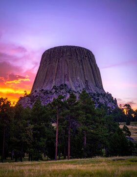 Devils Tower National Monument at sunset with vibrant sky and pine trees.