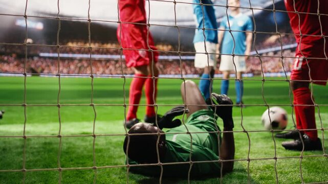 Defeated goalkeeper concedes goal as ball hits net on pitch, beaten by striker while successful opponents celebrate a winning goal in a stadium match.