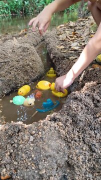 Medium angle shot of a child using hands to pick up and place plastic ducks and boats inside a hand-dug water trench next to a calm stream.