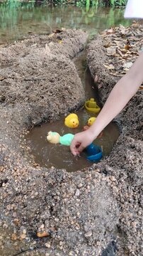 High-angle shot of a young child playing with colorful floating plastic toys inside a dug-out sand trench near a clear forest river during summer outdoor activity.