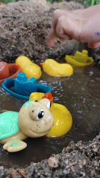 Macro close-up shot of colorful plastic duck and turtle toys floating in muddy water while a child plays in the background next to a riverbank.