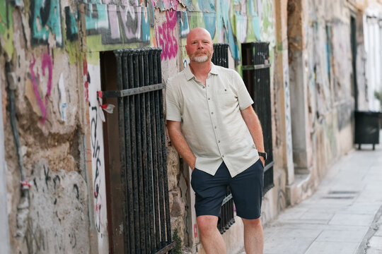 Man standing beside graffiti‑covered wall in Athens, Greece, wearing light shirt and navy shorts on a summer afternoon.