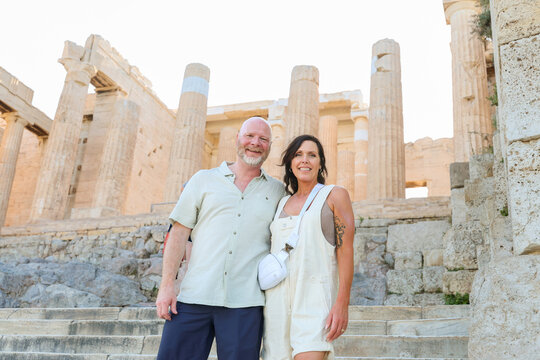 Smiling couple standing in front of ancient Greek temple at Acropolis in Athens, Greece, surrounded by tourists.